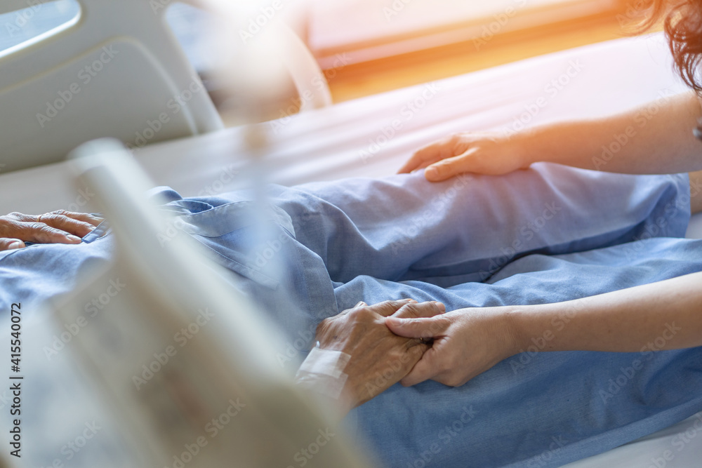 Fototapeta premium Caregiver and elderly senior patient (aged old adult person) holding hands in hospital bed or nursing hospice, geriatrician palliative home, while caretaker having medical health care service