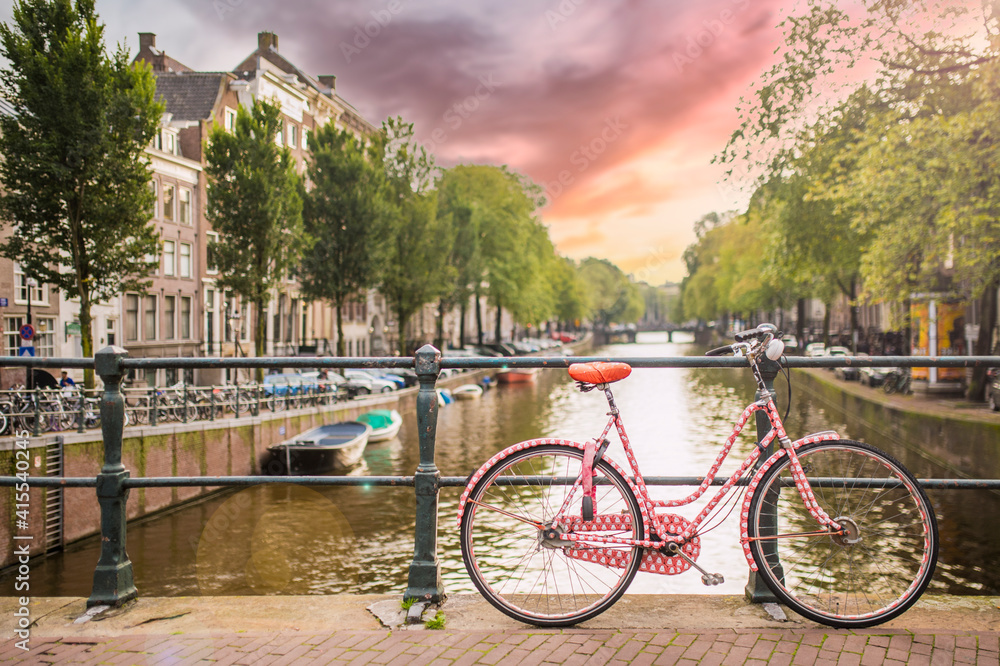 (Selective focus) Stunning view of a pink bicycle parked on a bridge that crosses one of Amsterdam's many canals. Beautiful sunset in the background.