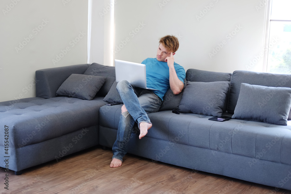 Young Caucasian man is thinking and searching online social media with laptop. He is sitting on sofa in living room. Work from home for Covod-19 outbreak. Lifestyle and techonology on holiday C Young Caucasian man is thinking and searching online social media with laptop. He is sitting on sofa in living room. Work from home for Covod-19 outbreak. Lifestyle and techonology on holiday C