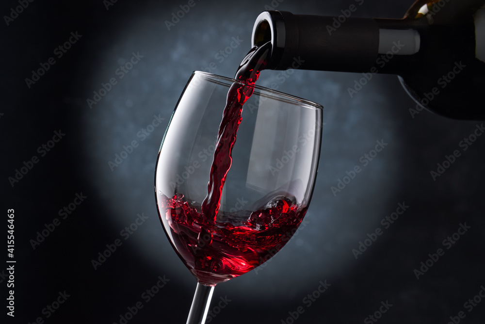 Fototapeta premium Red wine is poured into a glass from a bottle on a blurry blue background, a stream of red wine from the bottle swirls in the glass, close-up. Free space for text.