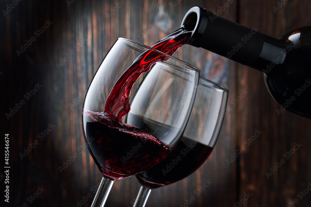 Fototapeta premium Red wine is poured into a glass from a bottle on a blurred wooden background, a stream of red wine from the bottle swirls in the glass, close-up. Free space for text.