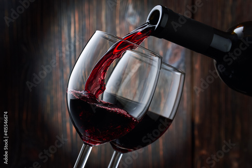 Red wine is poured into a glass from a bottle on a blurred wooden background  a stream of red wine from the bottle swirls in the glass  close-up. Free space for text.