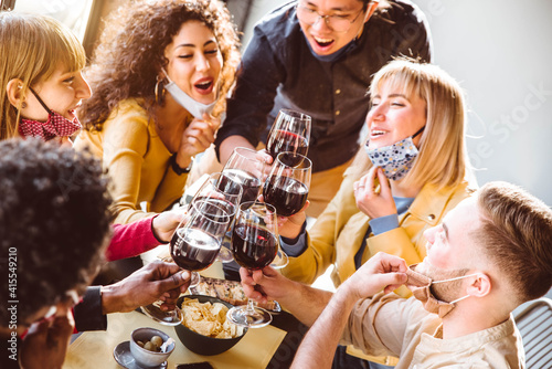 Happy friends wearing protective face masks toasting red wine sitting at restaurant - New normal friendship concept with young people having fun at home party - Focus on glasses