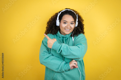 Young african american girl wearing gym clothes and using headphones smiling and pointing with hand and finger to the side © Irene