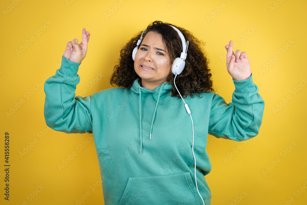 Young african american girl wearing gym clothes and using headphones gesturing finger crossed smiling with hope and looking side Young african american girl wearing gym clothes and using headphones gesturing finger crossed smiling with hope and looking side