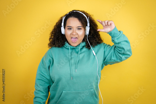 Young african american girl wearing gym clothes and using headphones smiling and confident gesturing with hand doing small size sign with fingers . Measure concept. © Irene