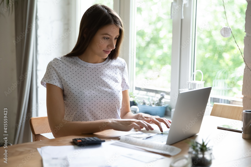 Millennial Caucasian woman sit at desk at home look at laptop screen manage household budget online. Happy young female work on computer calculate pay household expenses expenditures on web. Millennial Caucasian woman sit at desk at home look at laptop screen manage household budget online. Happy young female work on computer calculate pay household expenses expenditures on web.