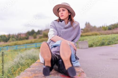 beautiful middle-aged young woman sitting on a stone wall in rural area wearing hat, gray pashmina, blue dress and black leather boots. spring summer fashion concept