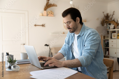 Serious millennial Caucasian businessman sit at desk at home office work online on laptop with documents. Pensive young man look at computer screen busy engaged in distant job, consulting client. © fizkes
