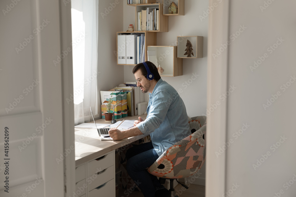 Candid view of millennial man in earphones sit at home office have webcam digital virtual meeting with colleagues on computer. Young male in headphones talk on video call on laptop with coworkers.