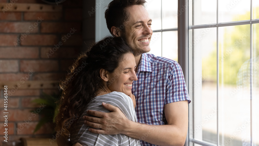 Close up smiling dreamy young couple hugging, visualizing good future together, happy beautiful woman and man dreaming, looking out window, standing at home and embracing, enjoying tender moment Close up smiling dreamy young couple hugging, visualizing good future together, happy beautiful woman and man dreaming, looking out window, standing at home and embracing, enjoying tender moment