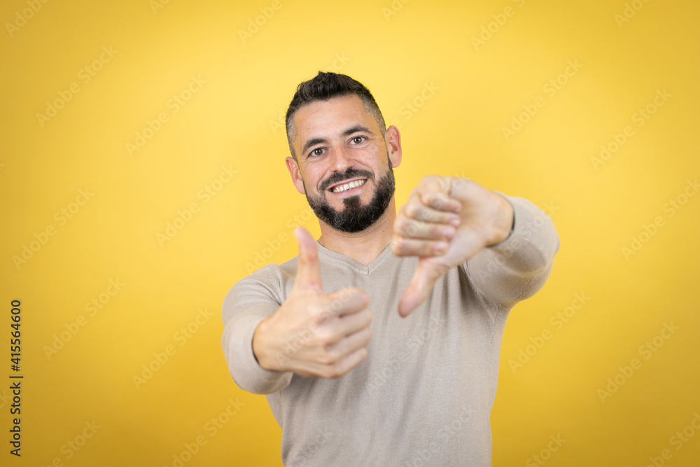Handsome man with beard wearing sweater over yellow background Doing thumbs up and down, disagreement and agreement expression. Crazy conflict