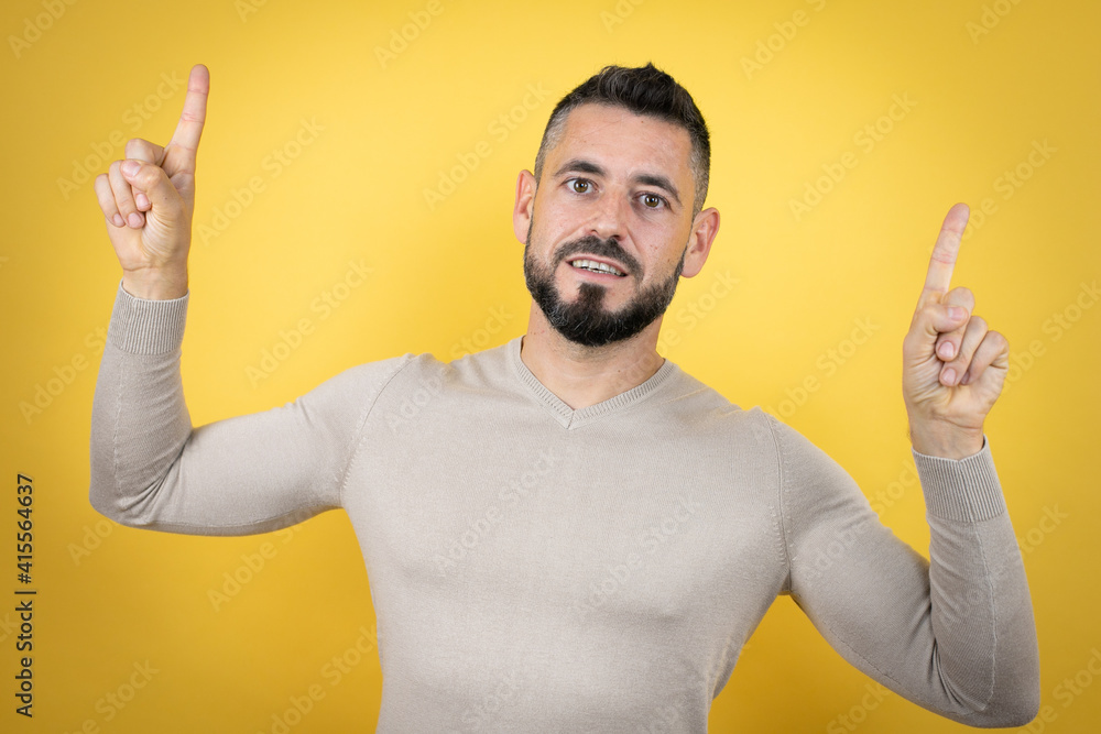 Handsome man with beard wearing sweater over yellow background smiling, looking at the camera and pointing up with fingers and raised arms Handsome man with beard wearing sweater over yellow background smiling, looking at the camera and pointing up with fingers and raised arms