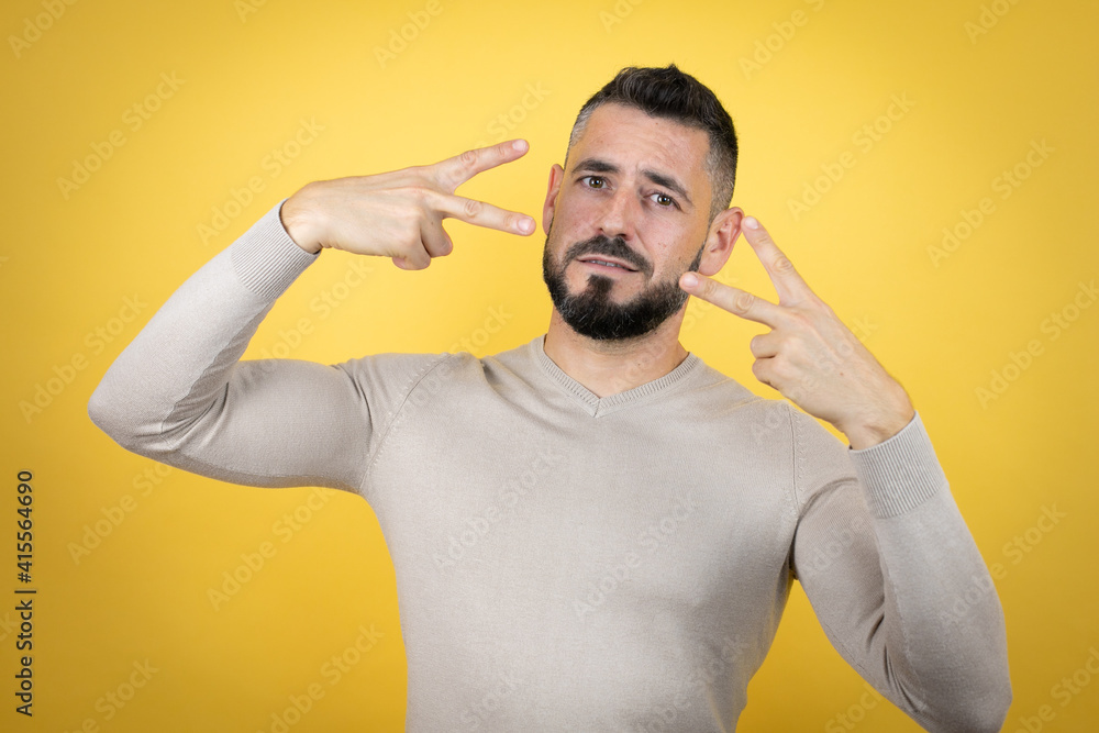 Handsome man with beard wearing sweater over yellow background Doing peace symbol with fingers over face, smiling cheerful showing victory Handsome man with beard wearing sweater over yellow background Doing peace symbol with fingers over face, smiling cheerful showing victory