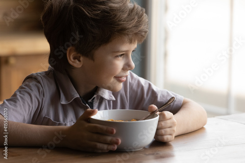 Close up smiling little boy holding bowl of tasty cereals with milk, happy adorable child kid enjoying breakfast in morning, starting new day, dreaming, sitting at wooden table in kitchen