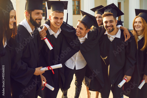 Group of smiling boys and girls students university graduates standing, hugging and celebrating getting diplomas with university building at background. Successful univesity graduation concept photo