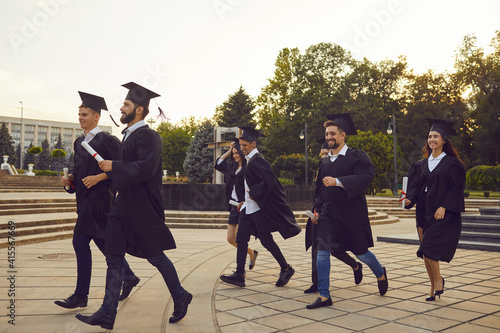 Group of young happy university graduates in traditional mantles walking with diplomas in hands outdoors and celebrating graduation. Successful univesity graduation concept photo