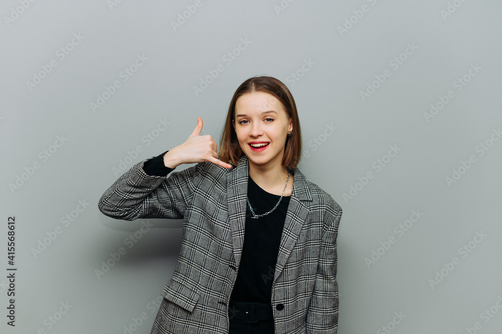 Positive woman in a jacket stands on a gray background, looks at the camera with a smile on his face and shows his fingers the gesture of the call. Positive woman in a jacket stands on a gray background, looks at the camera with a smile on his face and shows his fingers the gesture of the call.