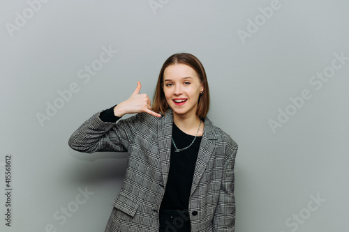 Positive woman in a jacket stands on a gray background, looks at the camera with a smile on his face and shows his fingers the gesture of the call.