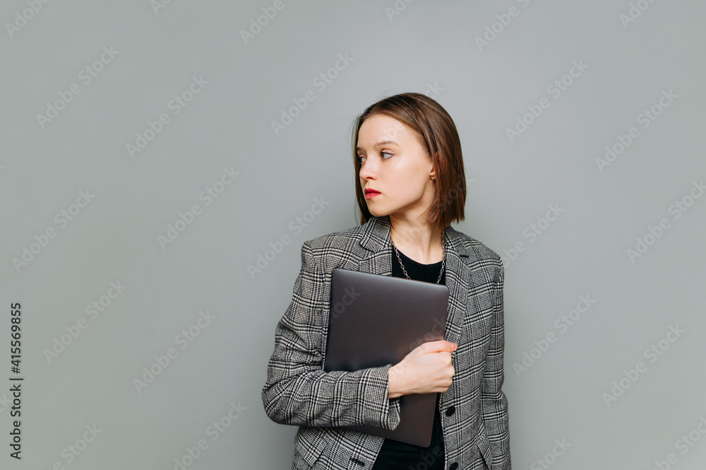 Attractive young business woman with a laptop in her hands stands on a background of a gray wall and looks to the side with a serious face wearing a jacket. Attractive young business woman with a laptop in her hands stands on a background of a gray wall and looks to the side with a serious face wearing a jacket.