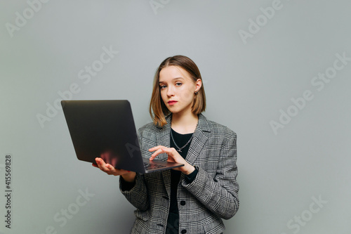 Woman freelancer in business. suit working on laptop on sirlmu background Stylish woman in smart casual clothes stands on a gray background with a laptop in hands and works, looks camera serious face