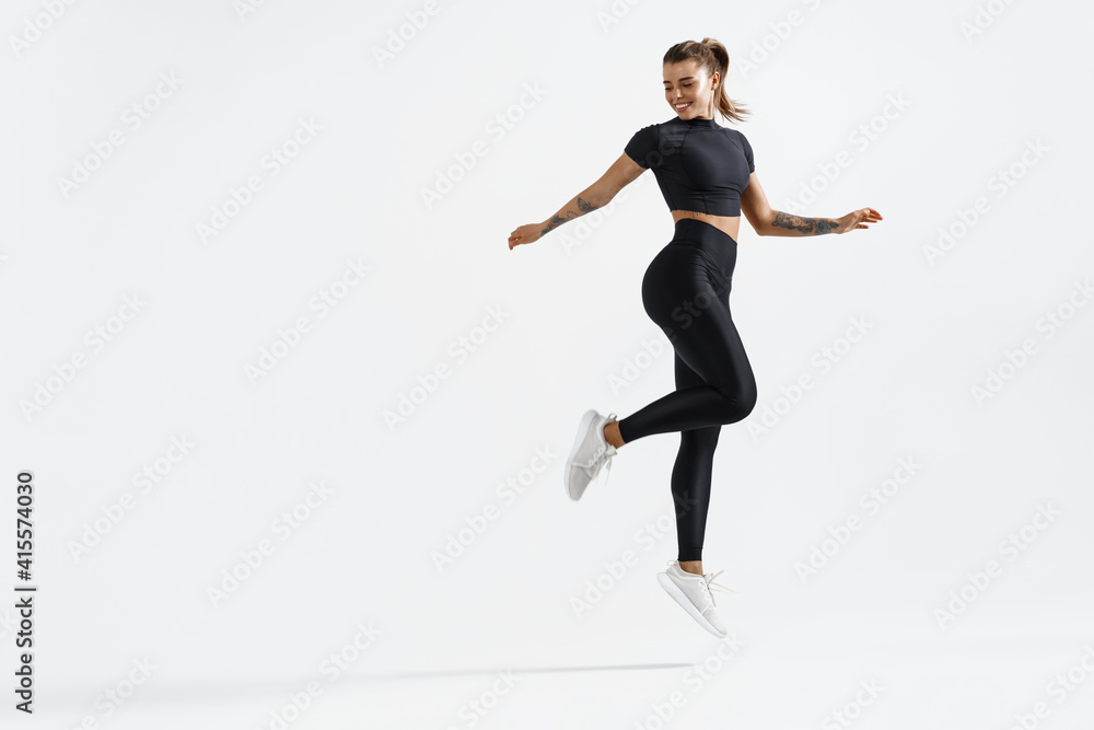 Fit and healthy sports woman runner jumping and looking behind. Female athlete in workout clothing doing exercises on white background, staring at empty space