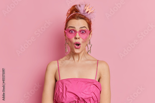 Studio shot of redhead fashionable woman stares surprisingly at camera keeps mouth opened reacts on amazing news wears trendy sunglasses and dress isolated over pink background. Style concept