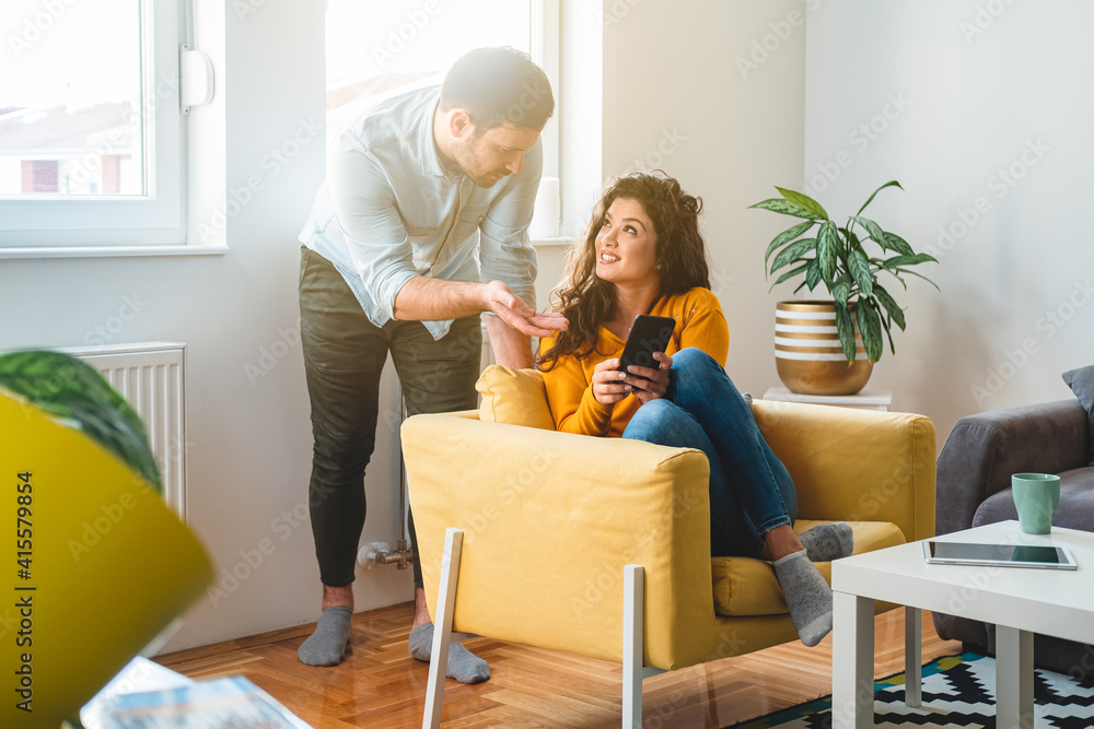 Smiling girlfriend showing mobile phone to serious boyfriend at home. Couple looking in smartphone together and talking in modern living room Smiling girlfriend showing mobile phone to serious boyfriend at home. Couple looking in smartphone together and talking in modern living room