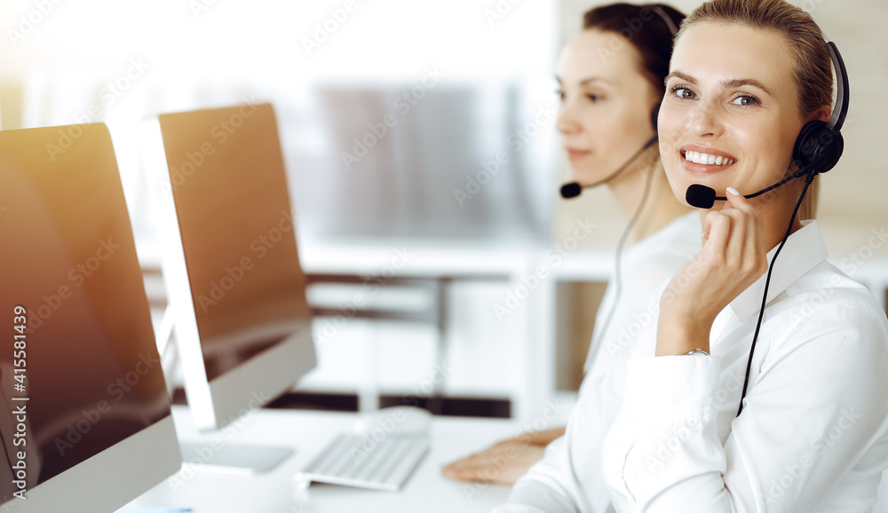 Blond female customer service representative is consulting clients online using headset in sunny office. Call center concept Blond female customer service representative is consulting clients online using headset in sunny office. Call center concept