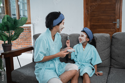 a mother and her little girl smile wearing a cream face mask wearing a towel and headband sitting on a chair