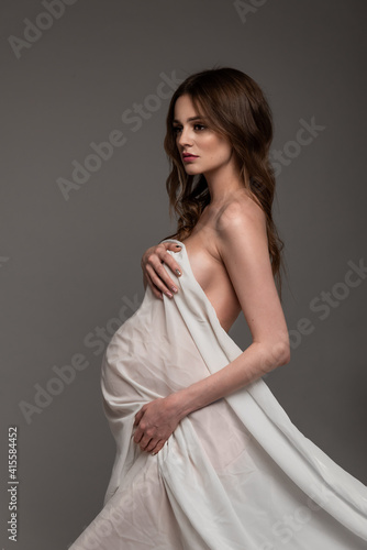 Pregnant girl in white flying cloth. Keeps belly and chest. Bare shoulder. Looking away. Grey background.