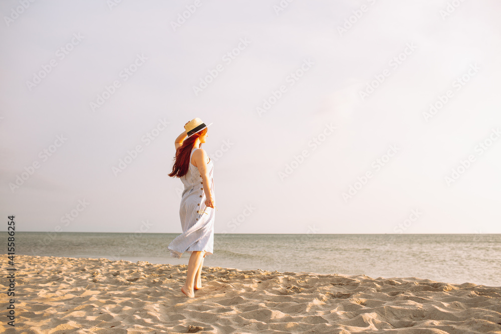 Young woman in dress with straw hat walk alone on empty sand beach at sunset sea shoreand and smiling.