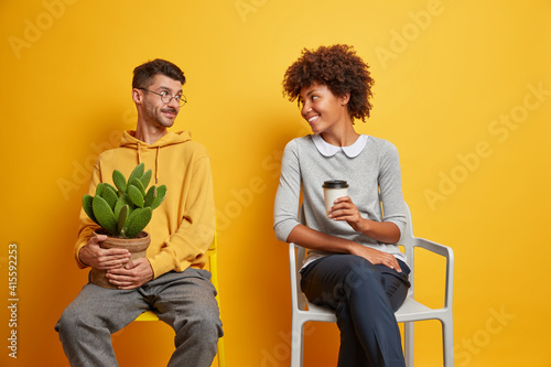 Cheerful woman and man have positive conversation with each other pose on chairs drink coffee get aquainted while waiting in queue isolated over yellow background. Keeping social distance concept photo