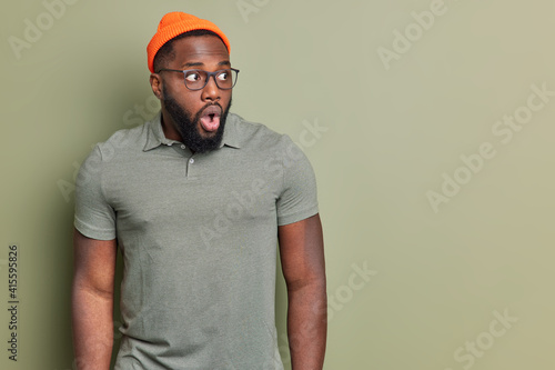 Portrait of shocked dark skinned man stares with widely opened eyes and mouth reacts emotionally on amazing news wears orange hat csual t shirt and spectacles isolated over green wal copy space photo