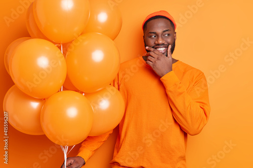 Positive dark skinned bearded man holds bunch of balloons celebrates festive occasion wears casual jumper and hat isolated over orange background being on party. Monochrome shot. Holidays concept © wayhome.studio 