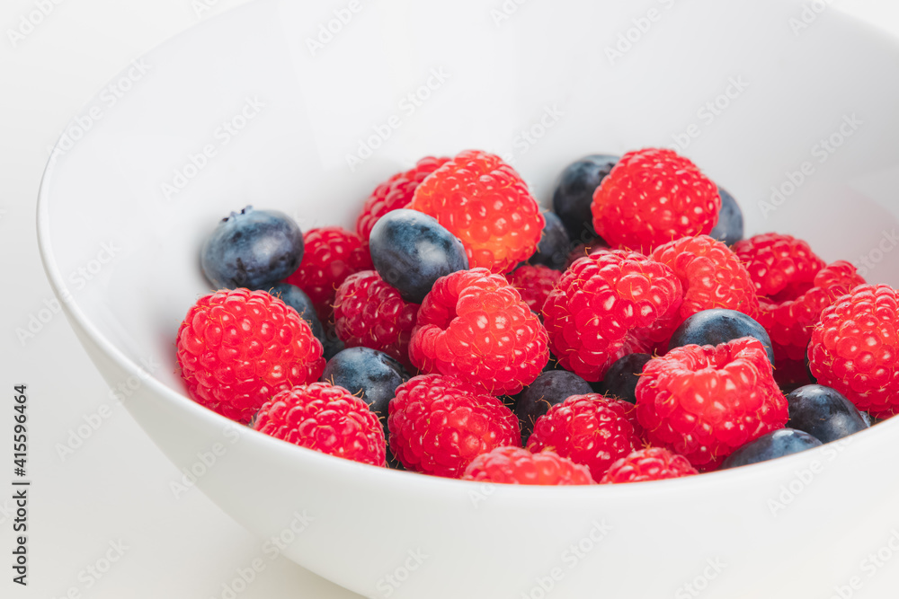 A close up detail of superfood mixed berries of raspberries and blueberries in a white bowl on a plain white surface and background. A close up detail of superfood mixed berries of raspberries and blueberries in a white bowl on a plain white surface and background.