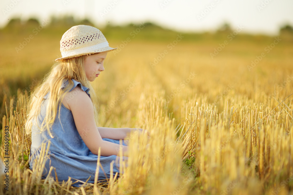 Obraz premium Adorable young girl having fun in a wheat field on a summer day. Child playing at hay bale field during harvest time. Kid enjoying warm sunset outdoors.