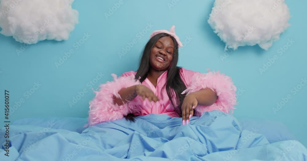 Energetic plump black woman with dark skin raises arms moves carefree dressed in domestic clothes enjoys serene atmosphere poses on bed applies collagen patches under eyes to reduce wrinkles Energetic plump black woman with dark skin raises arms moves carefree dressed in domestic clothes enjoys serene atmosphere poses on bed applies collagen patches under eyes to reduce wrinkles