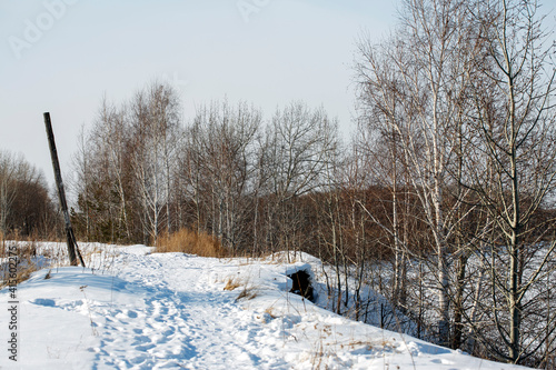 Snow-covered path in the village stretching into the distance. There is a cliff along the edge. A river flows at the bottom of the cliff. Winter landscape