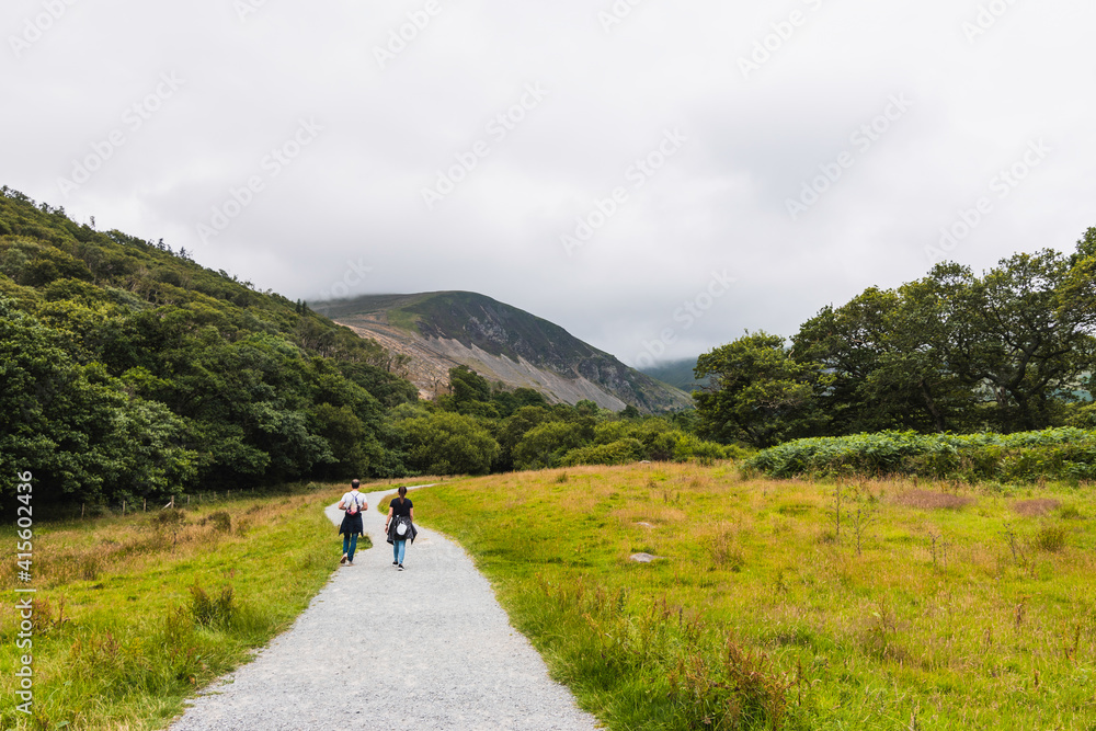 two people on their backs walking along a path in the direction of a mountain in a natural green landscape