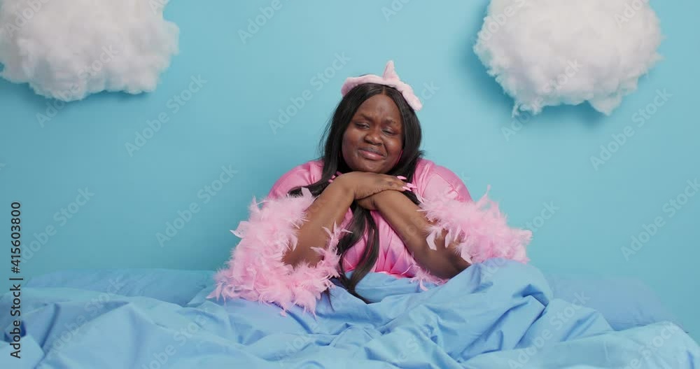 Frustrated black plump woman feels lonely experiences difficult moment looks stressed cries from despair poses in dressing gown under soft blanket isolated over blue background with white clouds Frustrated black plump woman feels lonely experiences difficult moment looks stressed cries from despair poses in dressing gown under soft blanket isolated over blue background with white clouds