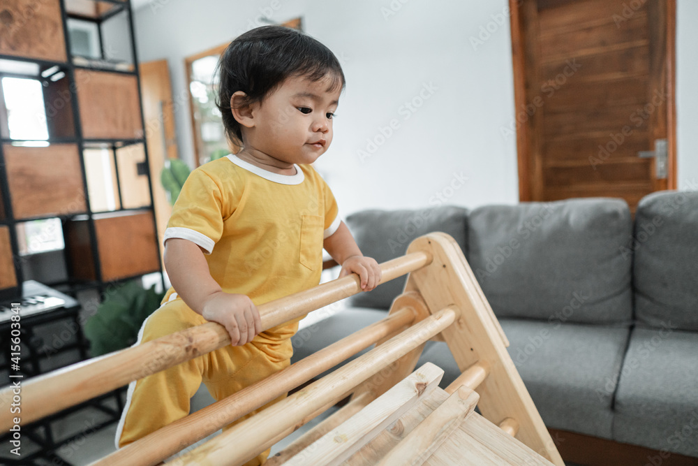 Fototapeta premium little kid looks down while climbing on the pikler triangle toy in the living room