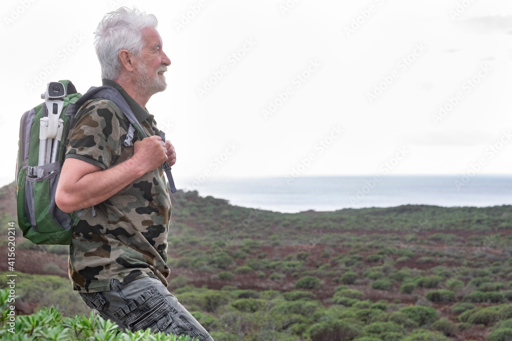 A smiling senior adult man with backpack on shoulders enjoying outdoors excursion between green bushes and sea. A white-haired elderly people in healthy activity
