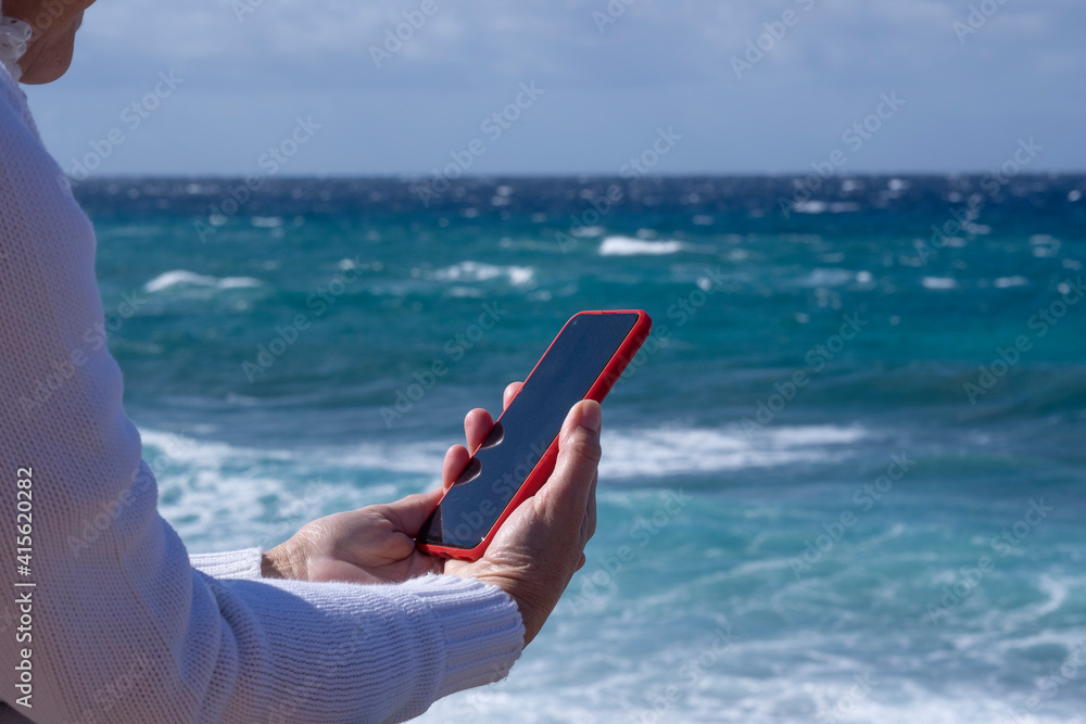A senior woman white dressed enjoying beach vacation on a windy day reading a message in her smartphone.  Relaxed smiling active retiree