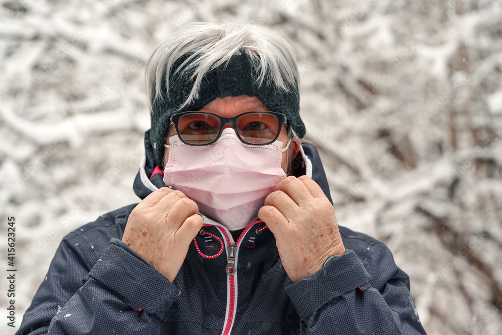Elderly senior woman with gray hair wearing pink disposable virus face mask, blurred winter snow covered trees background