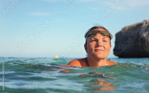 Young woman with swim goggles, surrounded by sea, closeup detail, rocky cliff and clear sky background © Lubo Ivanko