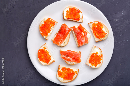 Bruschettes with butter and red caviar and salmon on a white plate on a gray concrete background.