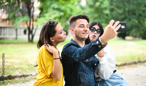 Cheerful smiling friends at the park sitting on a bench and taking selfies using a smart phone © Minerva Studio
