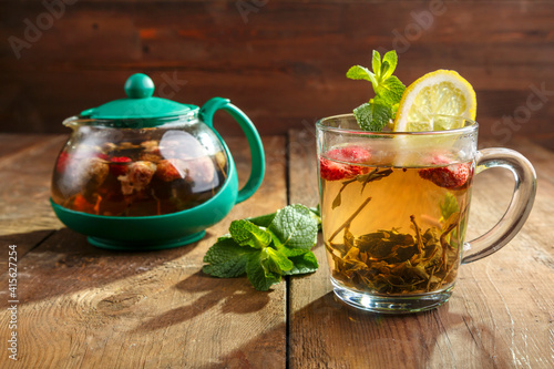 green tea in a glass cup with strawberries mint and lemon on a wooden table and a teapot and mint leaves.