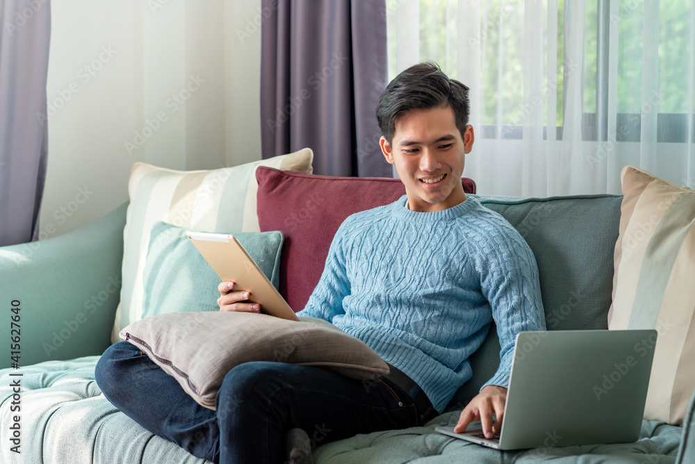 Young Asian man on a leisurely day sitting home on sofa using laptop and tablet on the couch. Young Asian man on a leisurely day sitting home on sofa using laptop and tablet on the couch.